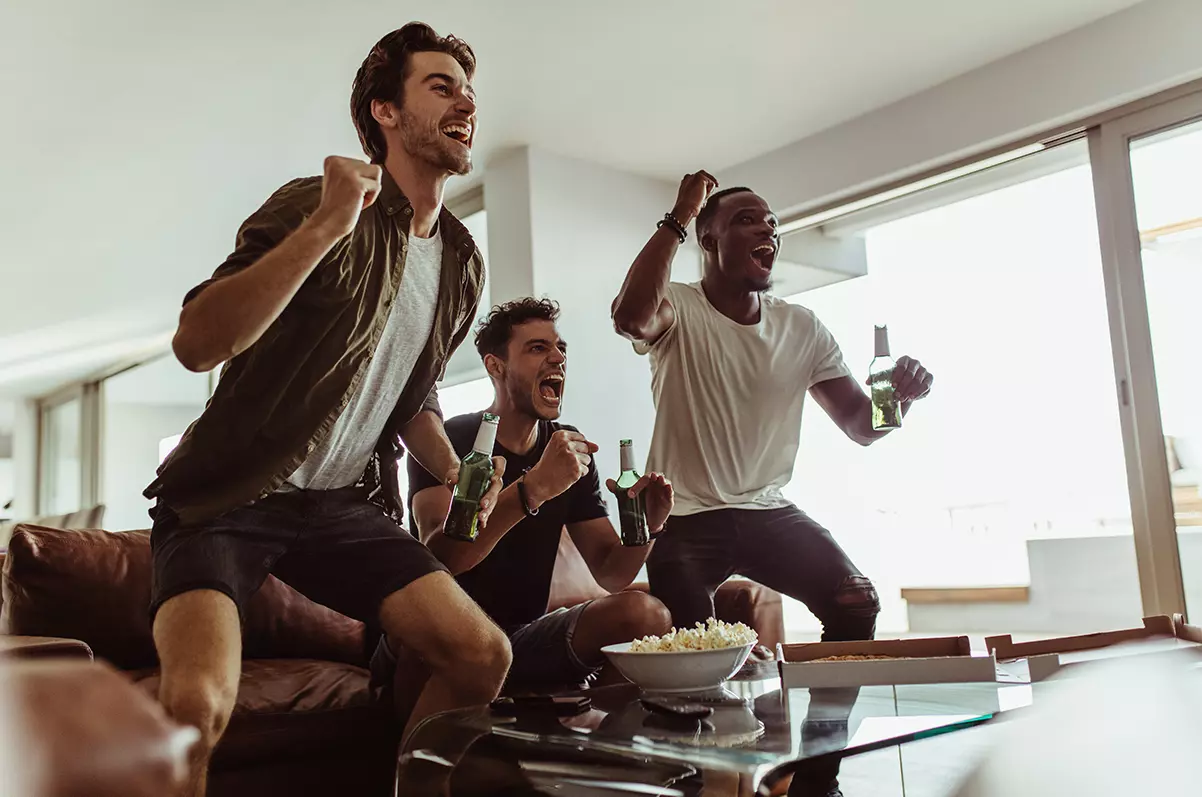 Three friends jumping in excitement holding beer bottles while watching sports game on TV.